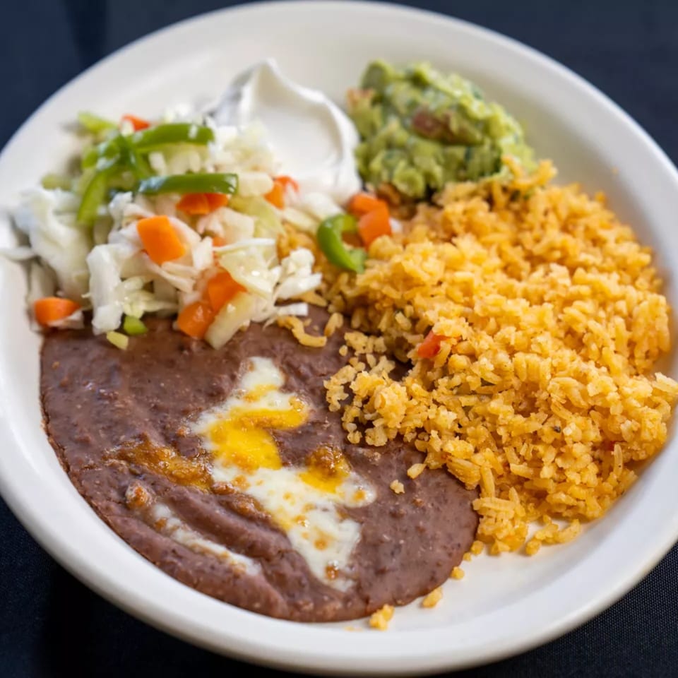 A white plate with refried beans, yellow rice, cabbage salad, sour cream, and guacamole.