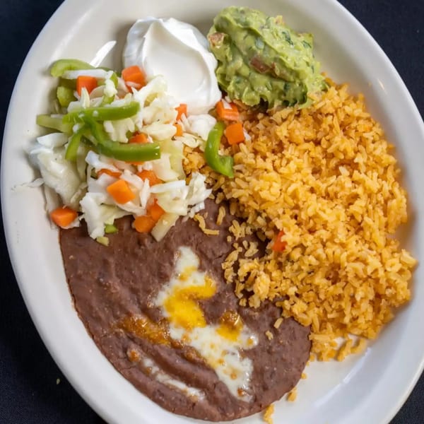 A white plate holds refried beans, Spanish rice, coleslaw, sour cream, and guacamole.