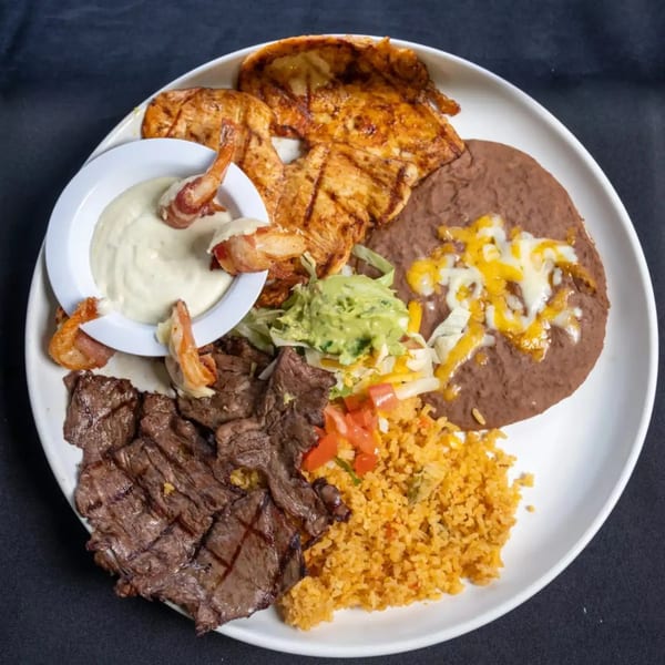 A large white plate filled with grilled chicken, steak, shrimp, rice, refried beans, and guacamole.